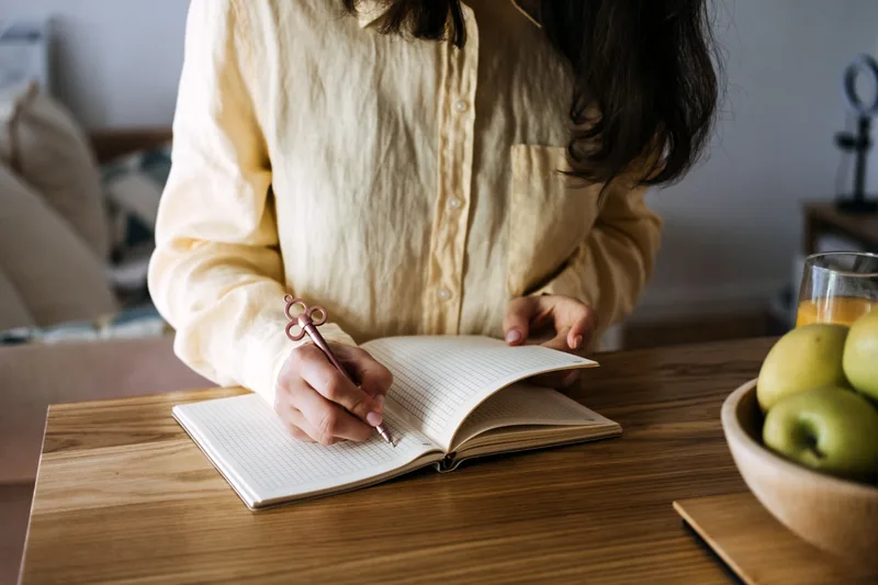 A woman writes in a notebook at a wooden table next to a bowl of green apples
