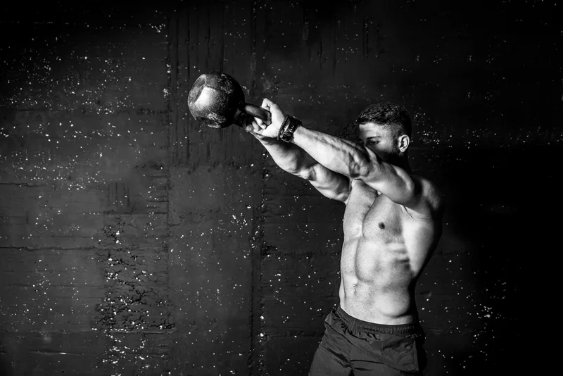 Athlete swinging a kettlebell overhead in a dark gym, photographed in dramatic black and white