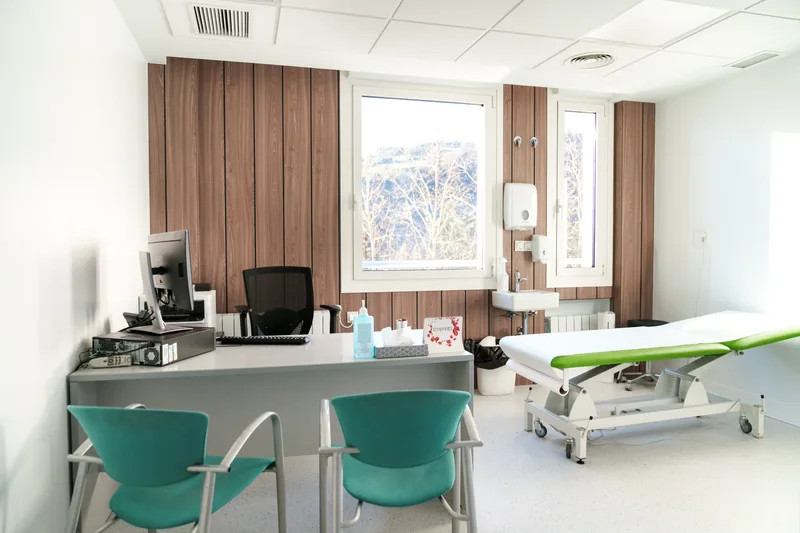 Clinic treatment room with a green-trimmed adjustable table, wood-panelled wall, and consultation desk