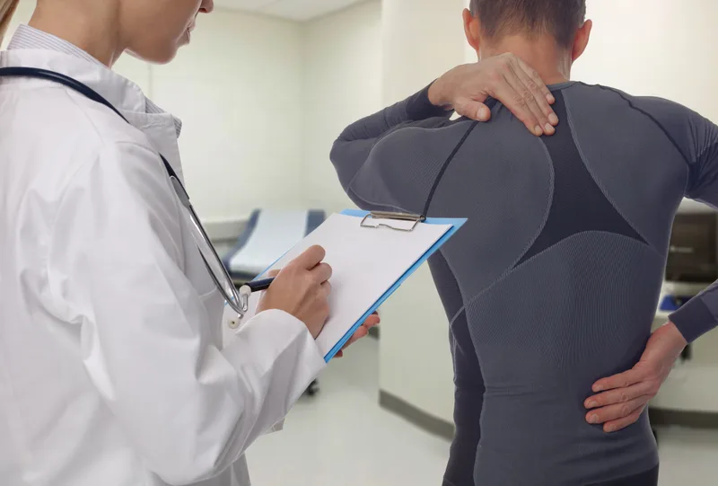 Clinician recording findings on a clipboard while assessing a patient's neck and upper back