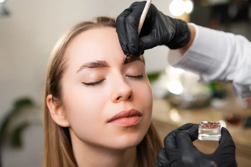 A gloved brow specialist brushing tint through a client's brows, eyes closed, dish of pigment in hand