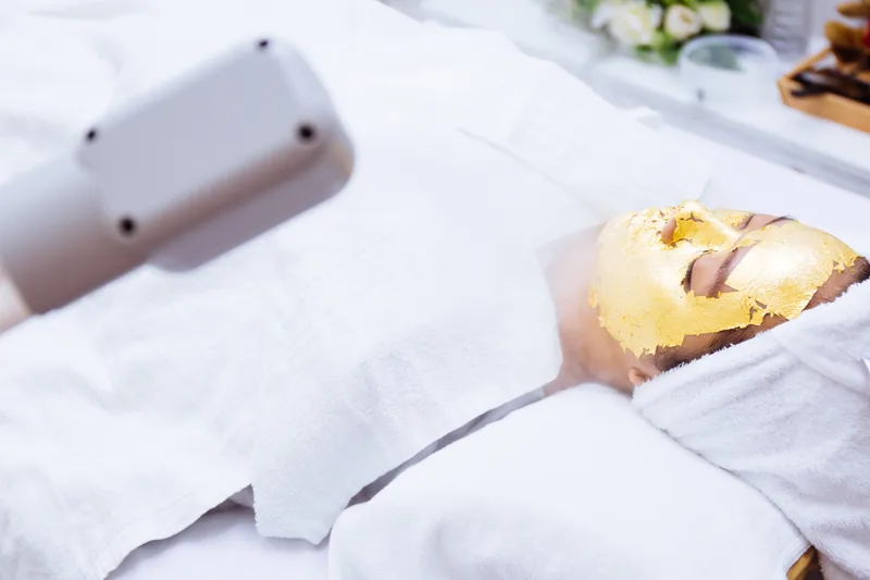 A client wrapped in white towels wearing a gold-leaf facial mask under warm treatment light
