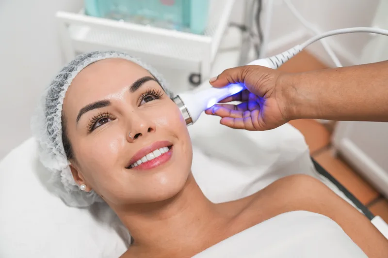An aesthetician gliding a glowing light-therapy handpiece across a relaxed client's cheek