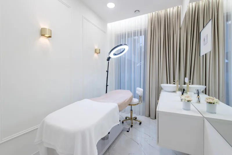 A Studio Lumière treatment room — white panelled walls, brass sconces, a ring light over the treatment bed, and marble floors