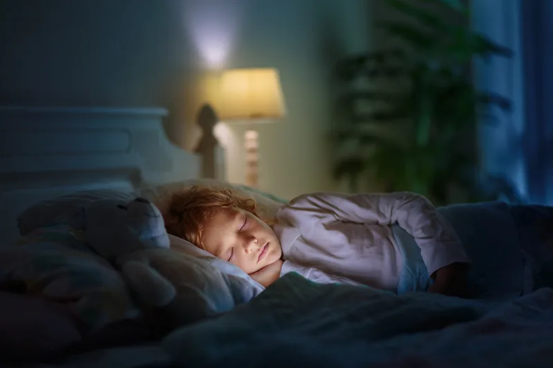A young child sleeping peacefully in bed beside a plush toy, lit by a warm bedside lamp