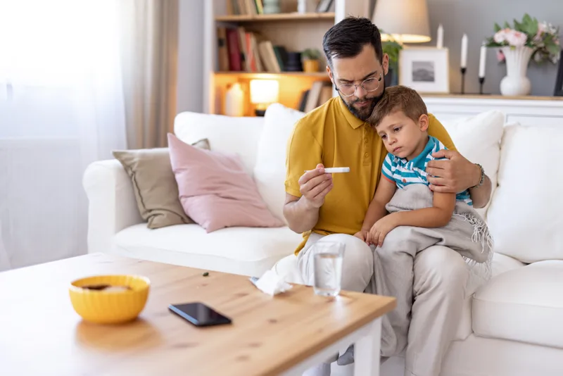 A father on the couch reading a thermometer while his young son rests against his arm