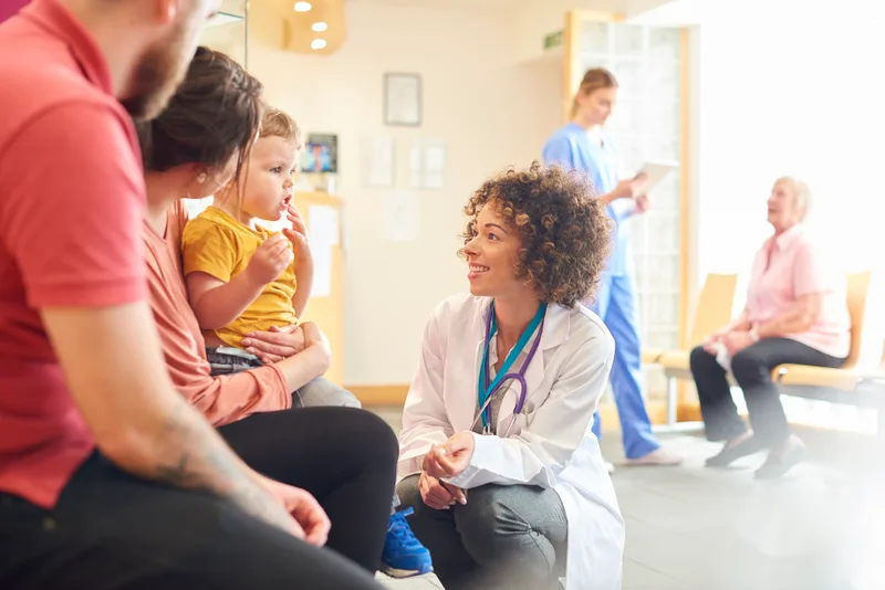 A pediatrician kneeling to chat with a toddler sitting on a parent's lap in the sunlit Westboro waiting room