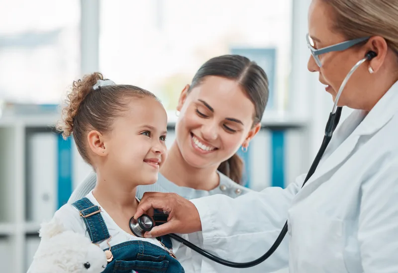 A pediatrician listening to a smiling young girl's chest with a stethoscope while her mother looks on