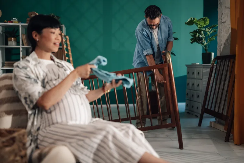 A pregnant woman holds tiny baby clothes while her partner builds a crib