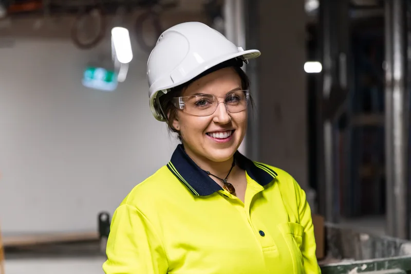 Mara Kovačić, master electrician, smiling in a white hard hat, safety glasses, and hi-vis shirt on a job site