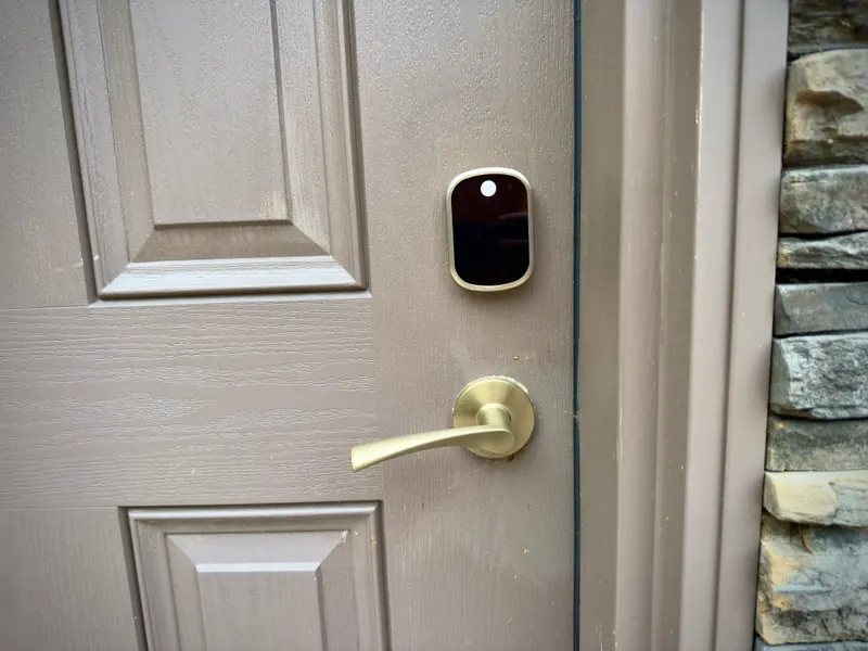 Brass keypad smart lock installed above the handle on a taupe front door beside a stone wall
