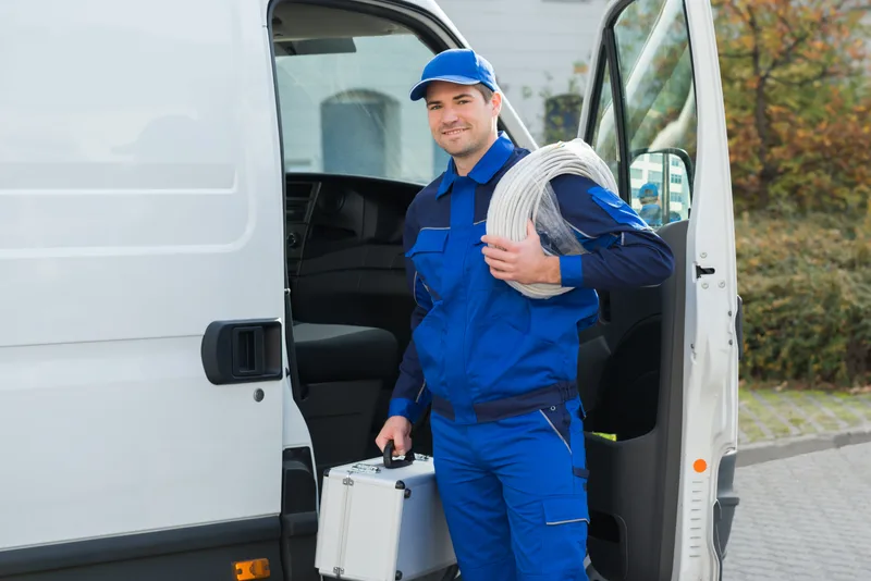 Cascade technician in a blue uniform and cap smiling beside his open service van, coil of supply line over one shoulder