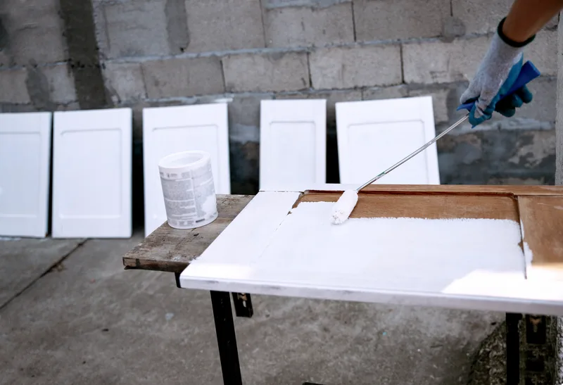 Cabinet doors lined up in the shop while a painter in gloves sprays white enamel on a door laid flat