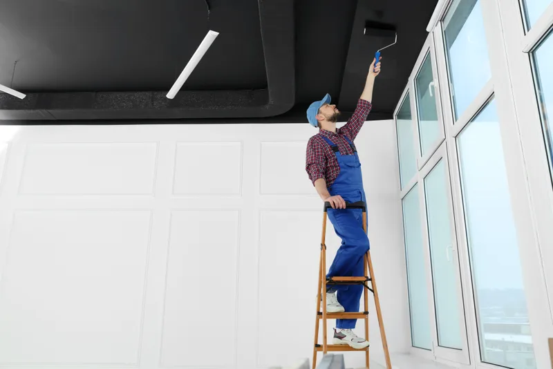Painter on a stepladder rolling the ceiling of a bright commercial space with tall windows