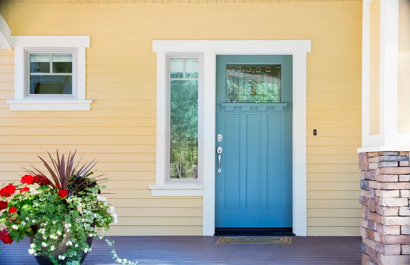 Teal front door with glass panes set into butter yellow siding with crisp white trim and a planter of red flowers