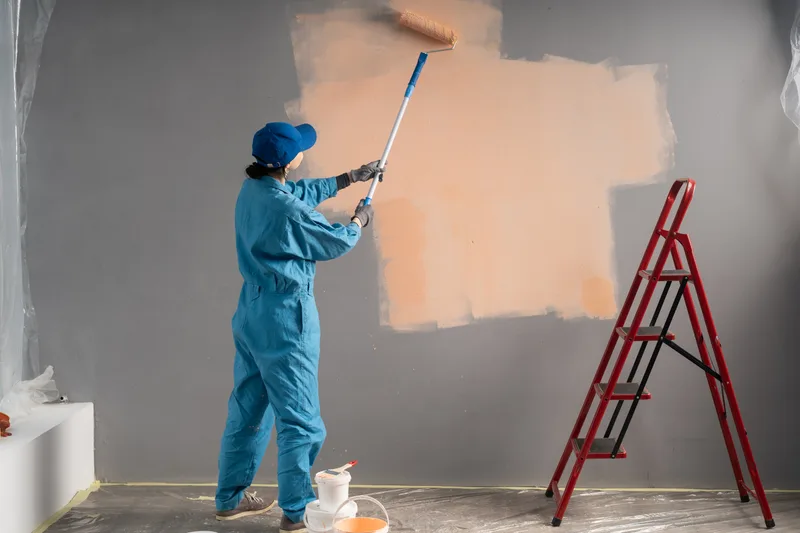 Cutline painter in blue coveralls rolling a warm peach colour over a grey wall with a long extension pole, red stepladder beside him