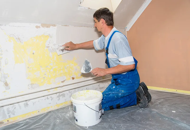 Painter kneeling on a plastic drop sheet, patching and smoothing a damaged wall with a putty knife before painting