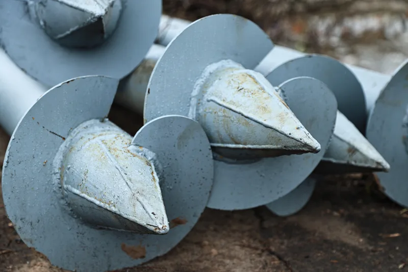Steel helical piers with spiral plates stacked on a job site, ready to be screwed into the ground