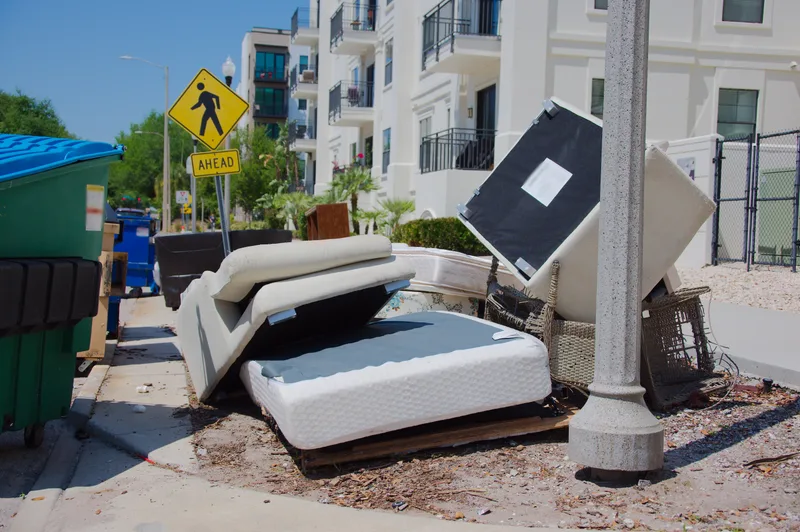 Mattresses, a folded sofa bed, and wicker furniture piled at a sunny city curb waiting for bulky-item pickup