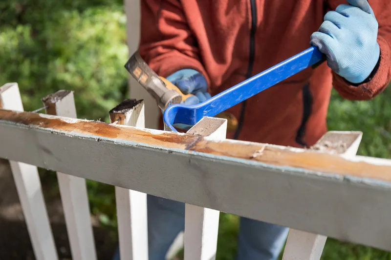 Worker in gloves prying apart a weathered white deck railing with a pry bar and hammer during a light demolition job