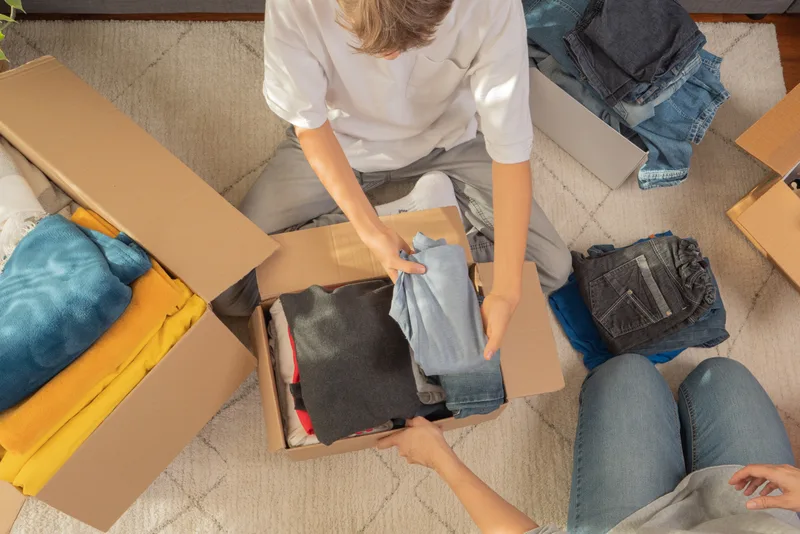 Overhead view of a family folding clothes and packing them into labeled cardboard donation boxes on a living room rug