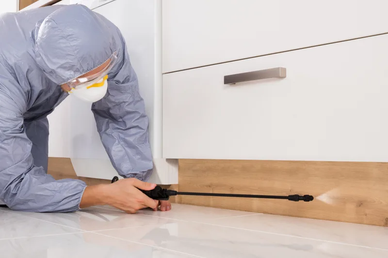 Technician in protective coveralls and goggles applying a precise crack-and-crevice treatment along a kitchen kickboard