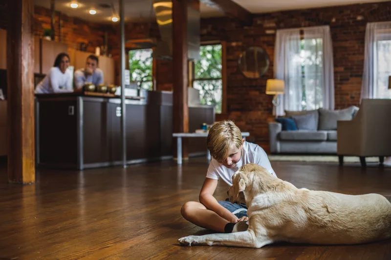 A boy sitting on a wooden living-room floor petting a yellow Labrador while his family watches from the kitchen