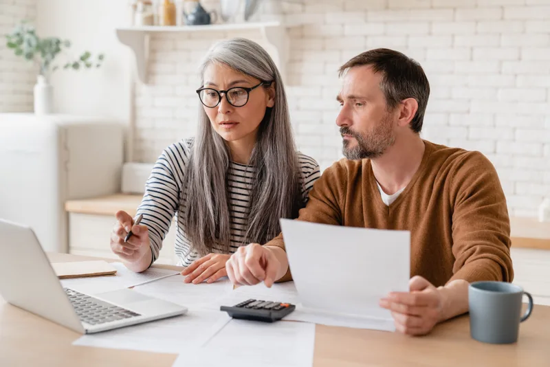 Couple at a kitchen table reviewing paperwork together with a laptop and calculator