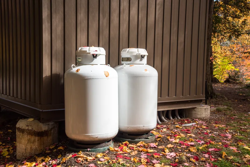 Two white propane cylinders set on the ground beside a building in autumn leaves