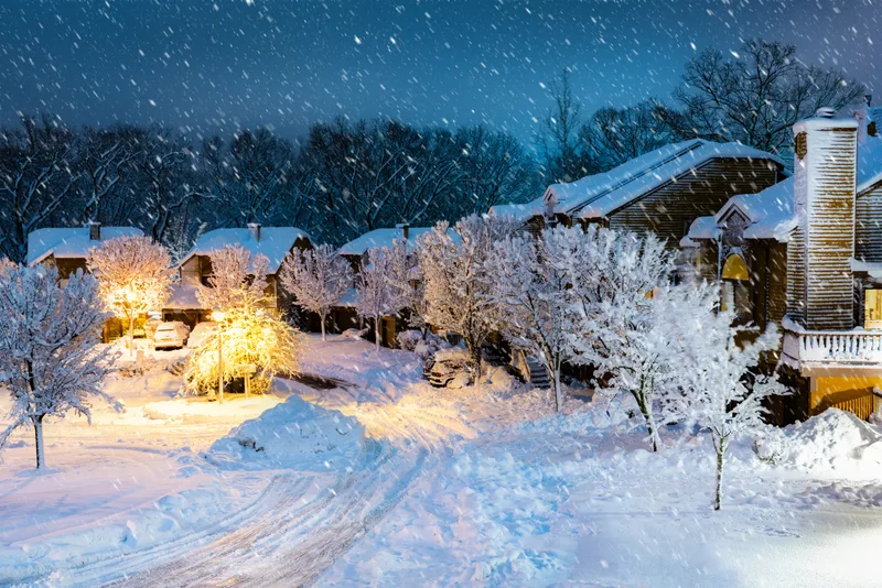 A row of houses with their windows and porch lights glowing during a heavy evening snowfall
