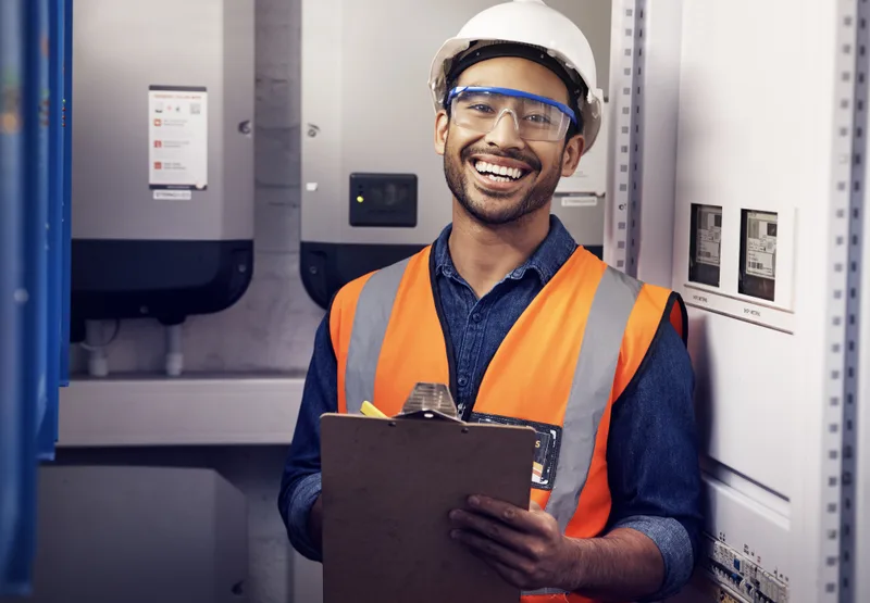 A smiling technician in a hard hat and safety vest holds a clipboard in an equipment room