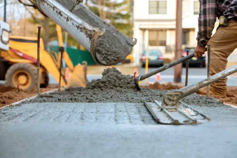 Concrete sliding down a truck chute into sidewalk forms while a finisher rakes it level, with a backhoe and houses behind