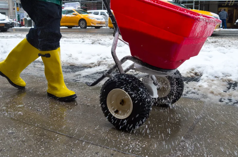 Worker in yellow boots pushing a salt spreader across an icy sidewalk