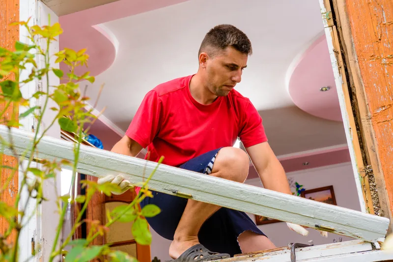Crew member kneeling in a bright room, easing an old window frame out of the wall
