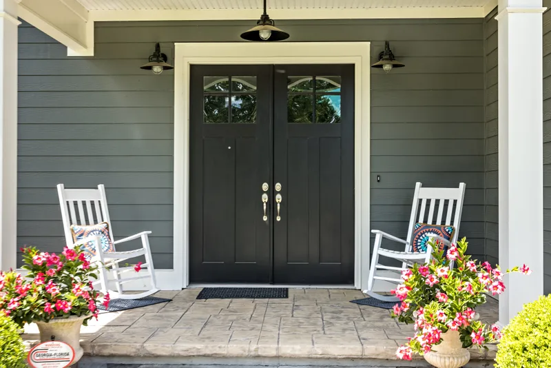 Black double entry doors with arched glass lites on a gray-sided porch with two white rocking chairs