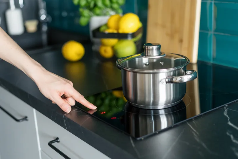 Hand touching the controls of a black glass cooktop with a steel pot on the burner