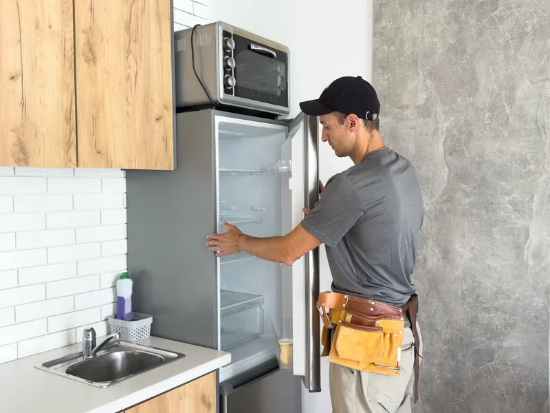 Technician in a cap and tool belt checking the inside of an open refrigerator in a bright kitchen