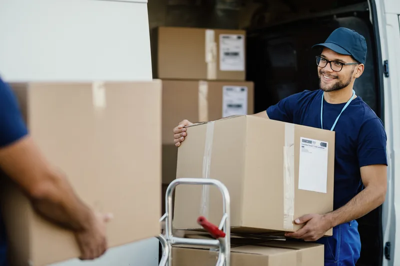 Smiling worker in a cap and glasses carrying a cardboard box out of the back of a van stacked with boxes