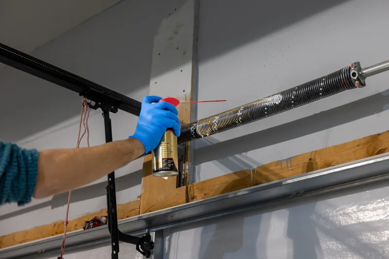 Gloved hand spraying lubricant along a garage door torsion spring during a tune-up