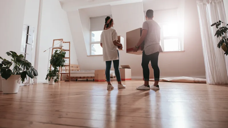 A couple carrying a packed cardboard box together across the wooden floor of a bright, nearly empty apartment