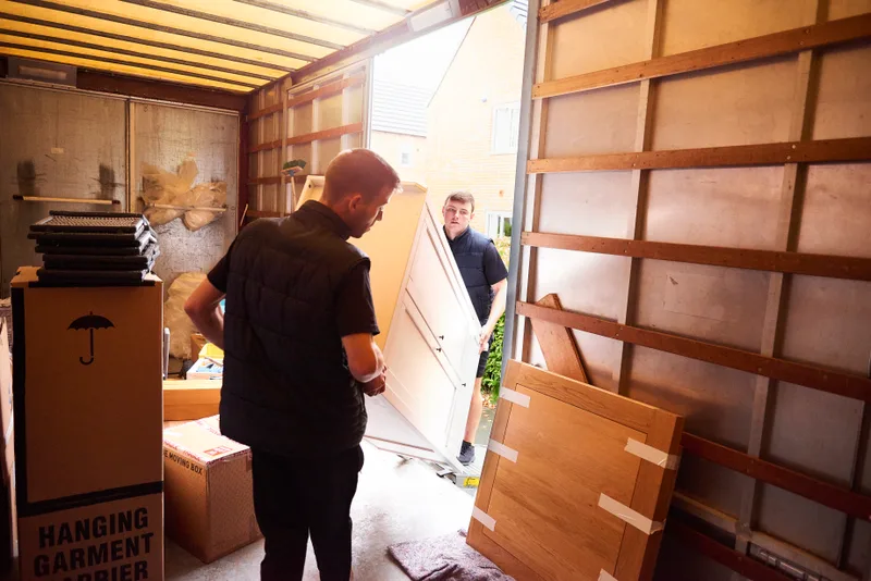 Two movers guiding a wardrobe panel into a padded moving truck stacked with labeled boxes and a garment carrier