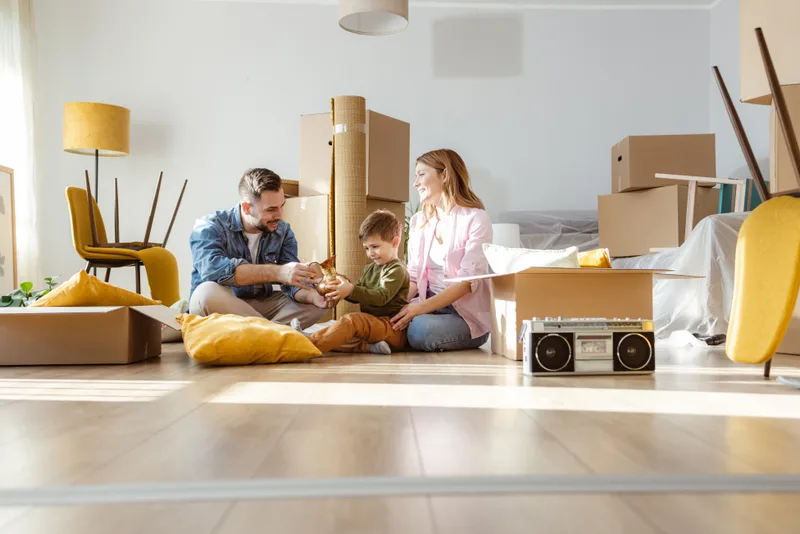 Parents and their young son unpacking a keepsake together on the floor of a sunlit new living room full of boxes