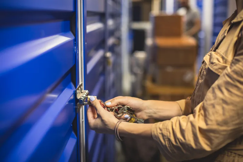 Hands turning a key in the lock of a blue roll-up storage unit door inside a clean storage facility corridor