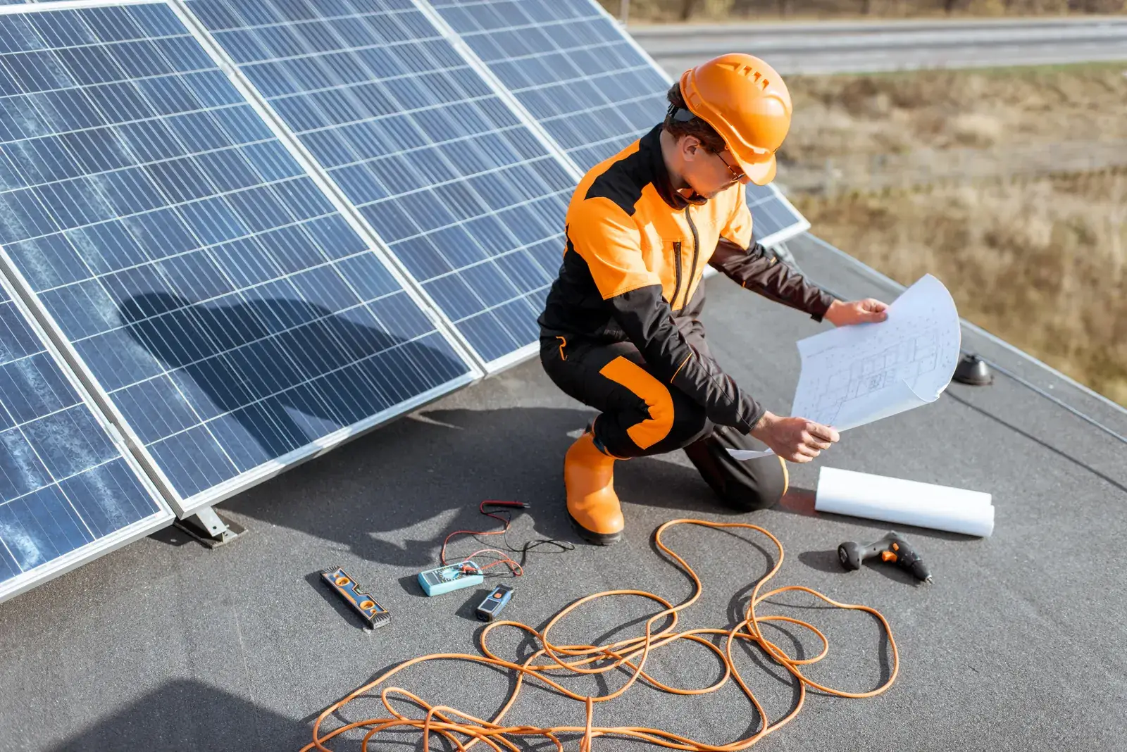 Solar installer in a safety harness reviewing layout drawings beside panels on a rooftop