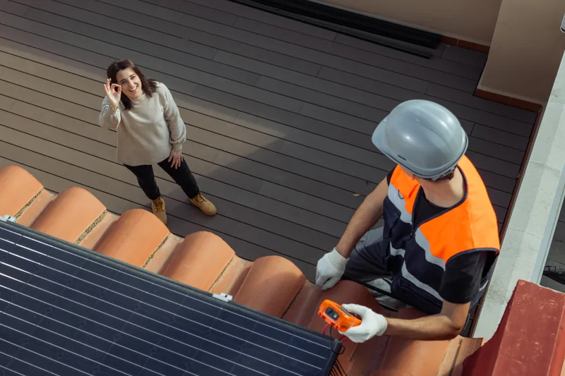 Technician in a hard hat testing a rooftop solar panel with a meter while the homeowner gives a thumbs-up from the patio