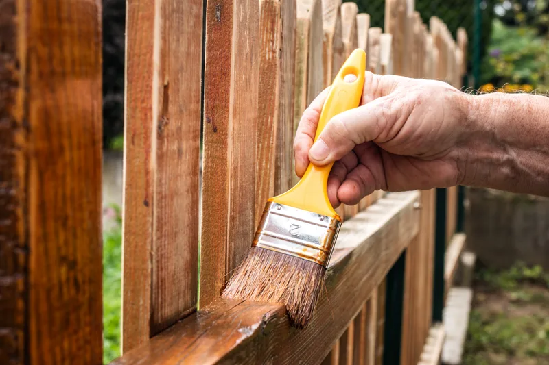 Brush applying stain to the rail of a wood picket fence