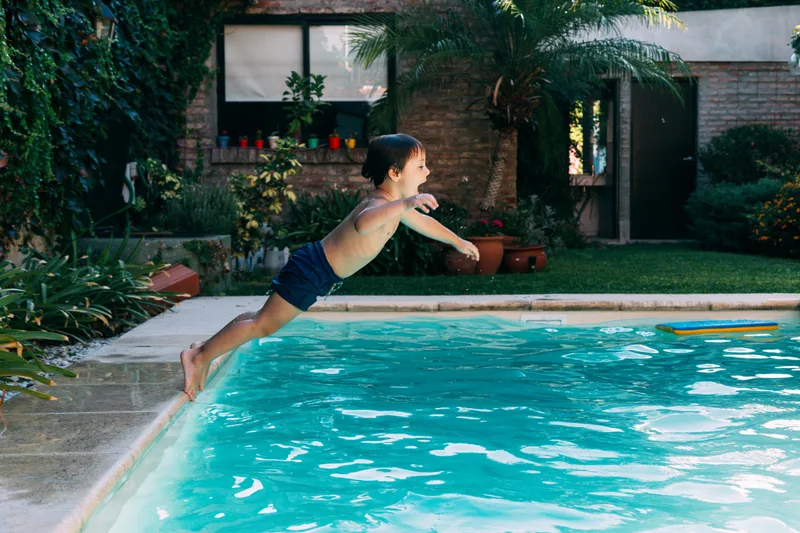 A young boy leaps into a bright blue backyard pool on a summer afternoon