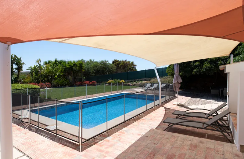 Backyard pool surrounded by a removable mesh safety fence, with a shade sail and loungers