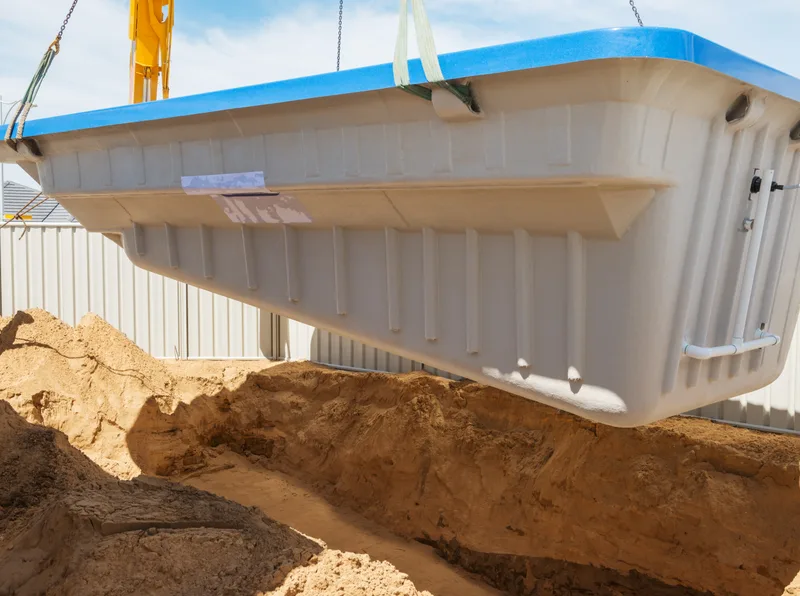 A one-piece fiberglass pool shell lifted by crane over the dug hole it will sit in