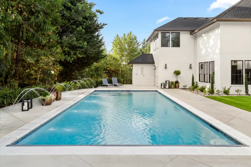 Rectangular gunite pool with deck jets arcing into the water, a white two-story house and a wide concrete deck